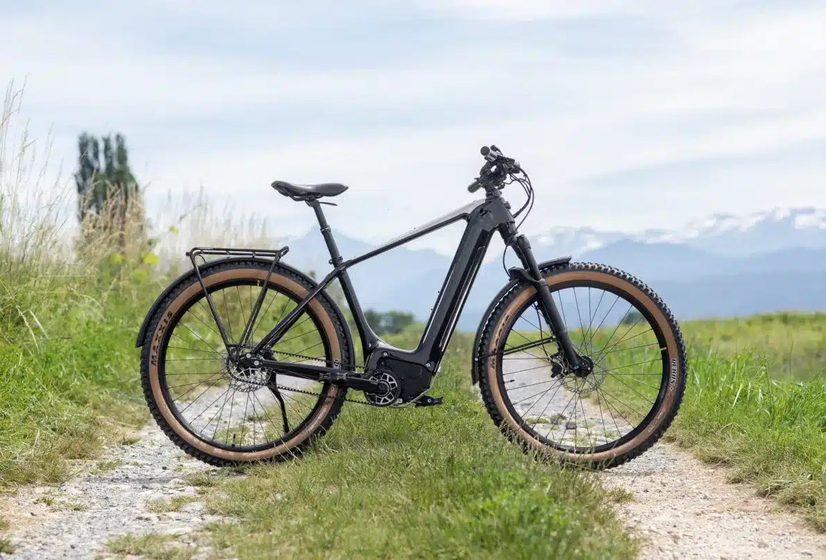 Schwarzes Elektro-Fahrrad 'Heritage Style Escapade' mit breiten Reifen und Gepäckträger, geparkt auf einem Feldweg mit Blick auf bergige Landschaften im Hintergrund.