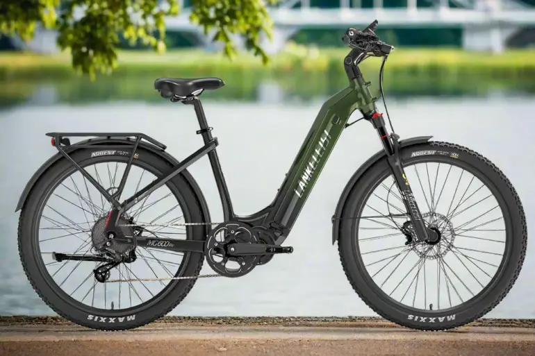 Grünes Lankeleisi MG600 Lite E-Trekkingbike mit breiten Reifen und Gepäckträger steht am Ufer, im Hintergrund ein Fluss und eine Brücke.