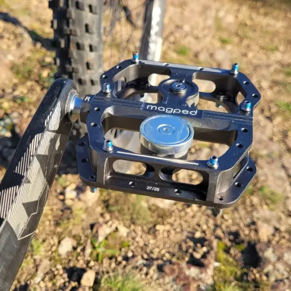 Nahaufnahme eines magped Enduro2 Fahrradpedals in Schwarz mit silbernem Magneten, montiert an einer Fahrradkurbel. Das Pedal zeigt blaue Schrauben und das Logo "magped".