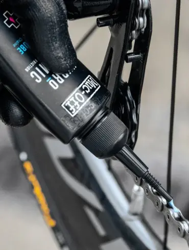 Nahaufnahme einer Hand mit schwarzem Handschuh, die Muc-Off Hydrodynamic Lube auf eine Fahrradkette aufträgt.