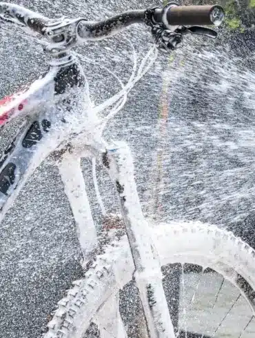 Ein Mountainbike wird mit dem Muc-Off Pressure Washer Bicycle Bundle gereinigt, wobei Schaum aufgetragen und mit Wasser abgespritzt wird. Deutlich erkennbar ist die pinke Flasche des Muc-Off Bike Cleaners, die am Hochdruckreiniger befestigt ist.