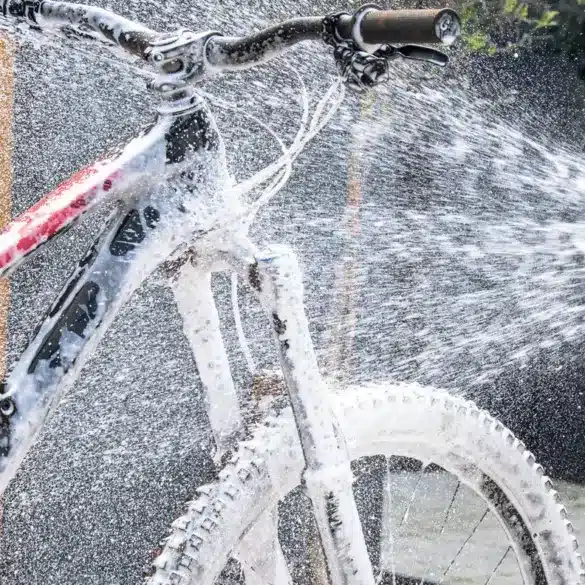 Ein Mountainbike wird mit dem Muc-Off Pressure Washer Bicycle Bundle gereinigt, wobei Schaum aufgetragen und mit Wasser abgespritzt wird. Deutlich erkennbar ist die pinke Flasche des Muc-Off Bike Cleaners, die am Hochdruckreiniger befestigt ist.