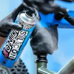 Hand mit schwarzem Handschuh sprüht Muc-Off Silicon Shine Spray auf den Rahmen eines Fahrrads, das in einem Montageständer eingespannt ist.