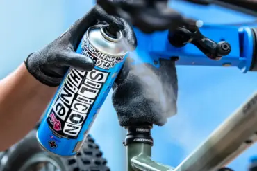 Hand mit schwarzem Handschuh sprüht Muc-Off Silicon Shine Spray auf den Rahmen eines Fahrrads, das in einem Montageständer eingespannt ist.