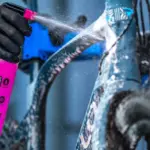 Eine Person mit schwarzen Handschuhen sprüht mit einer pinken Muc-Off Sprühflasche Reinigungsmittel auf ein blaues Fahrrad.