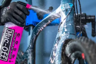 Eine Person mit schwarzen Handschuhen sprüht mit einer pinken Muc-Off Sprühflasche Reinigungsmittel auf ein blaues Fahrrad.