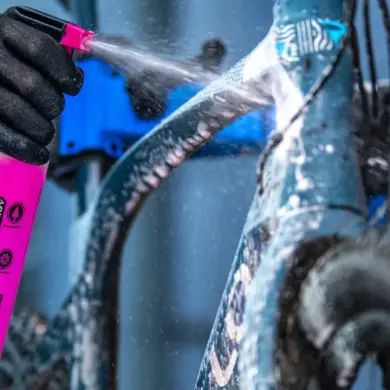 Eine Person mit schwarzen Handschuhen sprüht mit einer pinken Muc-Off Sprühflasche Reinigungsmittel auf ein blaues Fahrrad.