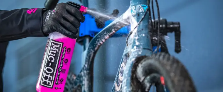 Eine Person mit schwarzen Handschuhen sprüht mit einer pinken Muc-Off Sprühflasche Reinigungsmittel auf ein blaues Fahrrad.