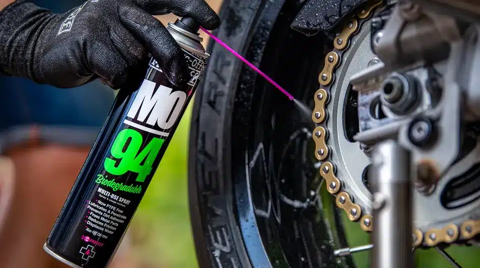 Nahaufnahme einer Hand mit schwarzer Handschuh, die eine Dose Muc-Off MO-94 Biodegradable Multi-Use Spray auf eine Fahrradkette sprüht.