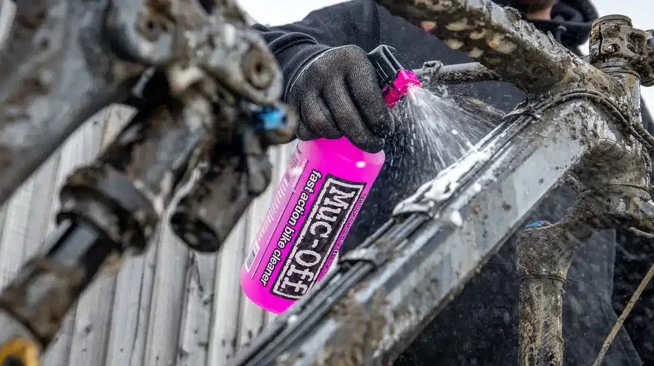 Person mit Handschuhen sprüht mit einer pinken Flasche Muc-Off Nano Tech Bike Cleaner einen stark verschmutzten Fahrradrahmen aus der Nähe ein.