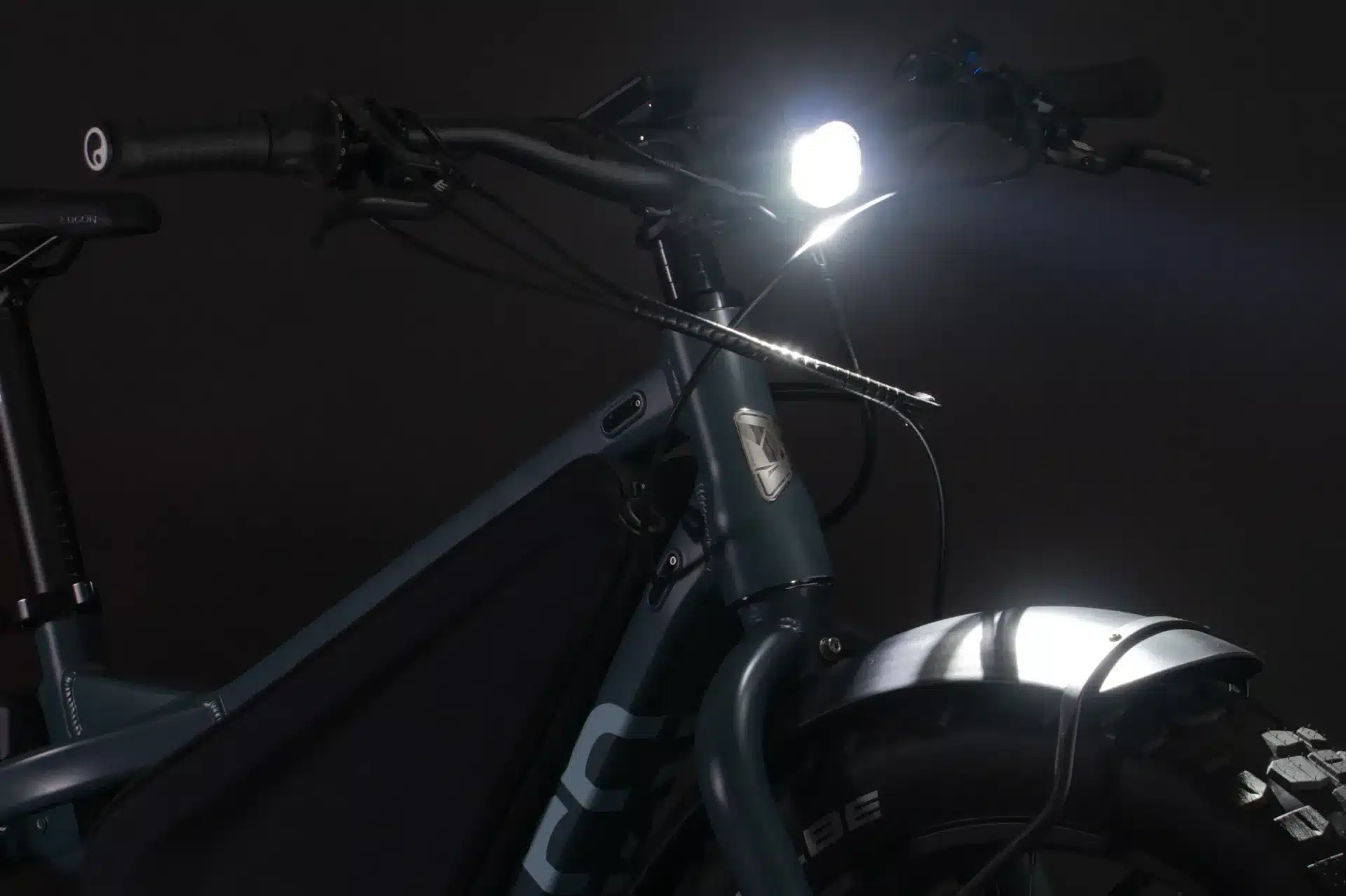 Nahaufnahme des vorderen Teils eines dunkelgrünen Tern Orox R14 Elektrofahrrads mit eingeschaltetem Scheinwerfer, der auf den schwarzen Lenker, die Federgabel und den breiten Vorderreifen mit Schlammschutzblech zielt.