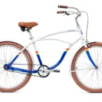 Ein weißes Priority Duzer Cruzer Fahrrad mit einem Traverse-Rahmen, braunem Sattel und Griffen, braunen Reifen, Schutzblechen und blau-gelb-roten Streifen auf dem Rahmen. Das Fahrrad ist auf einem weißen Hintergrund isoliert.