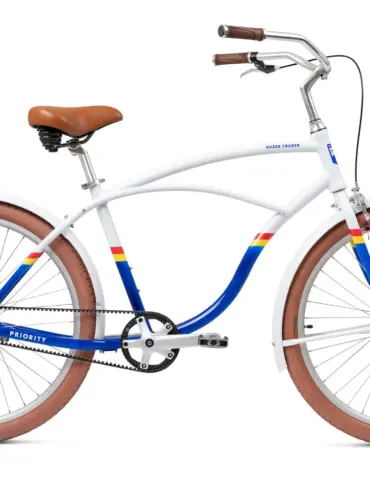 Ein weißes Priority Duzer Cruzer Fahrrad mit einem Traverse-Rahmen, braunem Sattel und Griffen, braunen Reifen, Schutzblechen und blau-gelb-roten Streifen auf dem Rahmen. Das Fahrrad ist auf einem weißen Hintergrund isoliert.