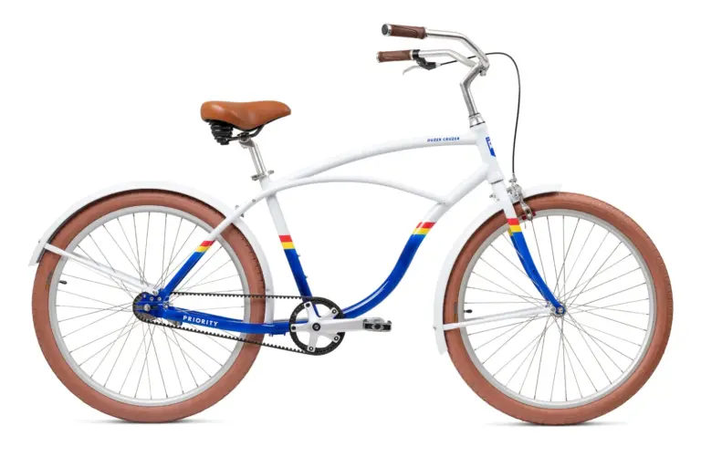 Ein weißes Priority Duzer Cruzer Fahrrad mit einem Traverse-Rahmen, braunem Sattel und Griffen, braunen Reifen, Schutzblechen und blau-gelb-roten Streifen auf dem Rahmen. Das Fahrrad ist auf einem weißen Hintergrund isoliert.