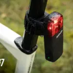 Das Sigma RECO 81 LINK, ein smartes Radar-Rücklicht, ist an der Sattelstütze eines Fahrrads montiert. Das rote Rücklicht leuchtet und die schwarze Einheit zeigt das Sigma-Logo. Im Hintergrund ist eine verschwommene grüne Naturkulisse zu sehen. Ein IP67-Schutzsymbol ist links unten platziert.