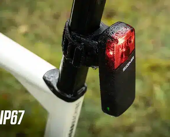 Das Sigma RECO 81 LINK, ein smartes Radar-Rücklicht, ist an der Sattelstütze eines Fahrrads montiert. Das rote Rücklicht leuchtet und die schwarze Einheit zeigt das Sigma-Logo. Im Hintergrund ist eine verschwommene grüne Naturkulisse zu sehen. Ein IP67-Schutzsymbol ist links unten platziert.