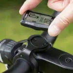 Ein Finger hält das SIGMA EOX VIEW 700 E-Bike Display, das an einer Fahrradlenkerhalterung befestigt ist. Das Display zeigt verschiedene Fahrdaten und die Uhrzeit an.