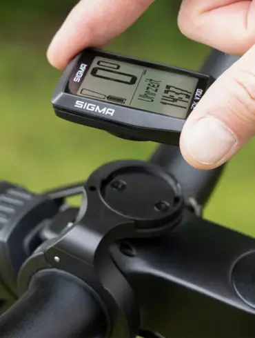 Ein Finger hält das SIGMA EOX VIEW 700 E-Bike Display, das an einer Fahrradlenkerhalterung befestigt ist. Das Display zeigt verschiedene Fahrdaten und die Uhrzeit an.