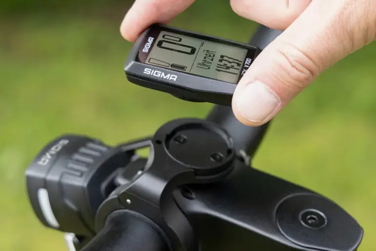 Ein Finger hält das SIGMA EOX VIEW 700 E-Bike Display, das an einer Fahrradlenkerhalterung befestigt ist. Das Display zeigt verschiedene Fahrdaten und die Uhrzeit an.