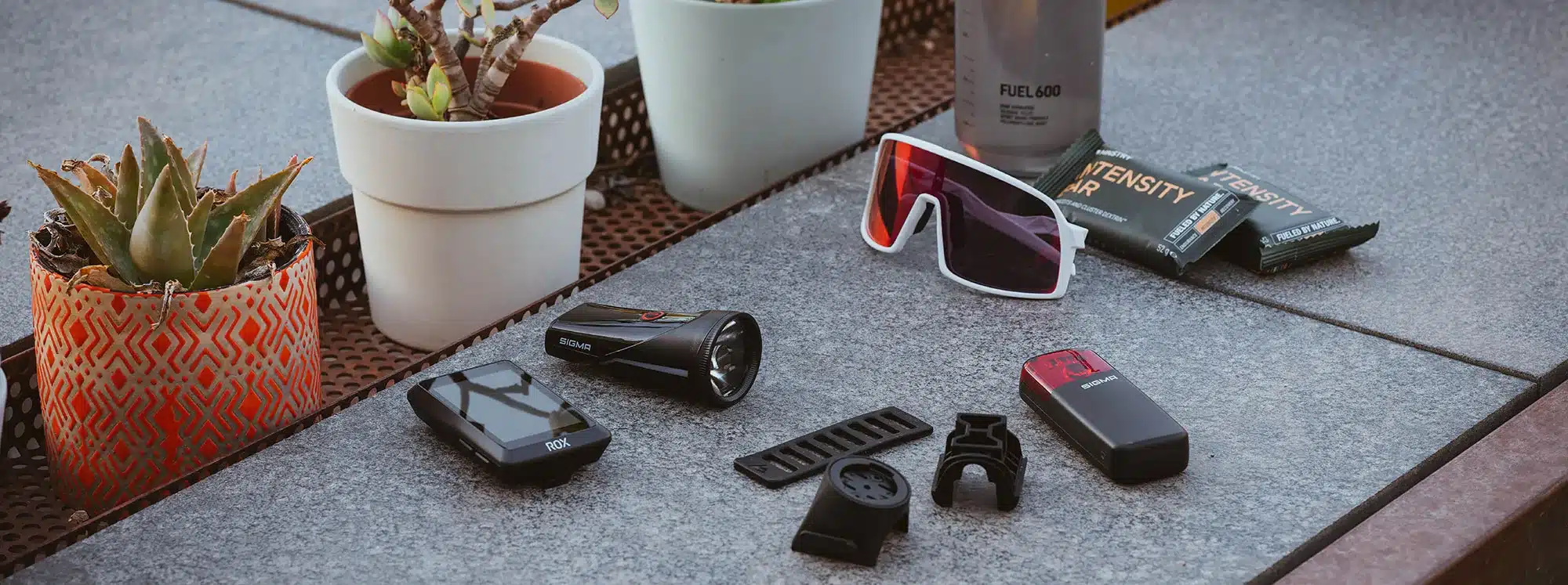 Produktfoto mit SIGMA ROX Fahrradcomputer, SIGMA RECO 80 LINK Rücklicht, Montagezubehör, Sportbrille und Energieriegeln auf einer grauen Oberfläche.