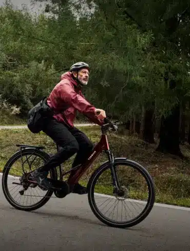Mann mit Helm und Umhängetasche fährt ein dunkelrotes TENWAYS Ago Air E-Bike auf einer Kurve einer asphaltierten Straße durch einen Wald mit hohen Bäumen.