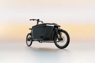 Ein schwarzes TENWAYS CARGO ONE Lastenrad mit breiter Ladebox und integrierter Beleuchtung, steht auf einem neutralen, hellen Untergrund.