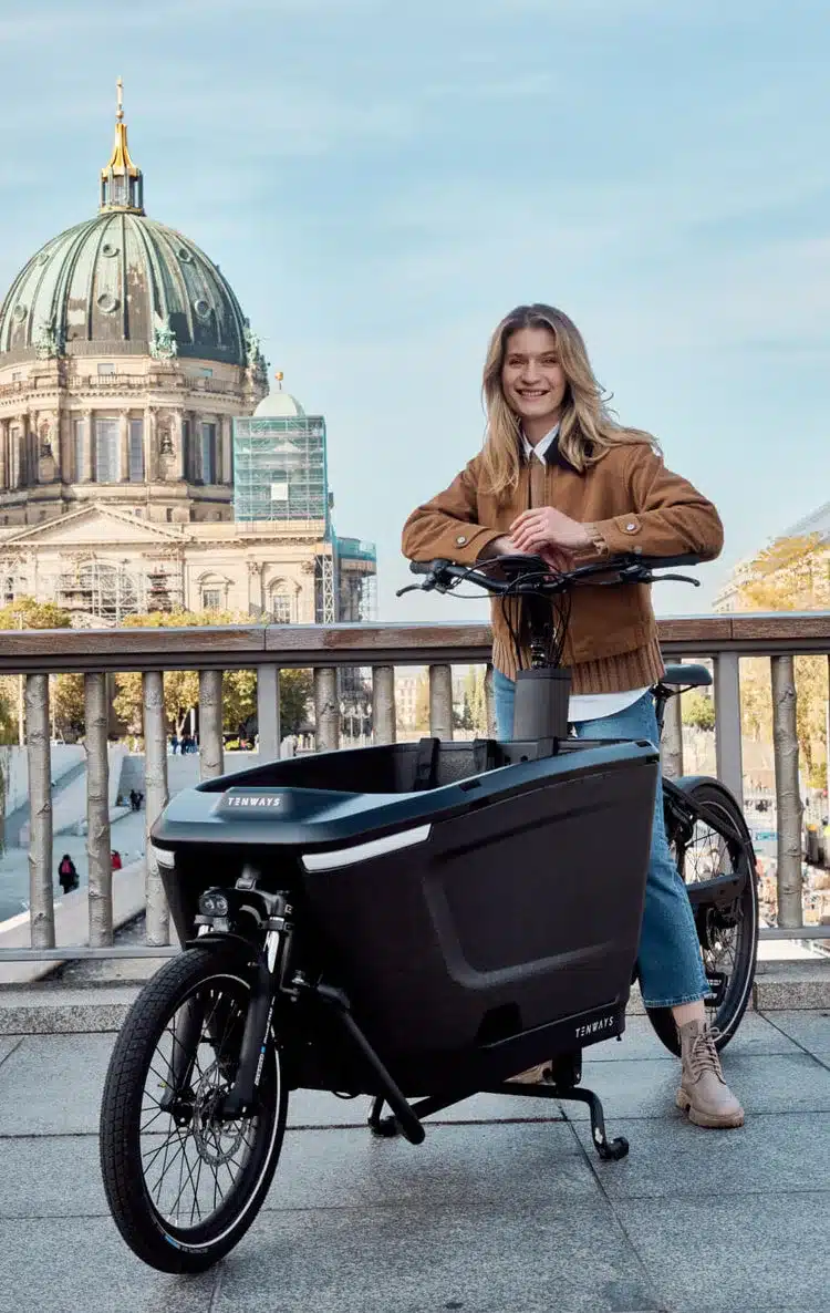 Das schwarze TENWAYS CARGO ONE Lastenrad mit einer jungen Frau davor, mit dem Berliner Dom im Hintergrund.