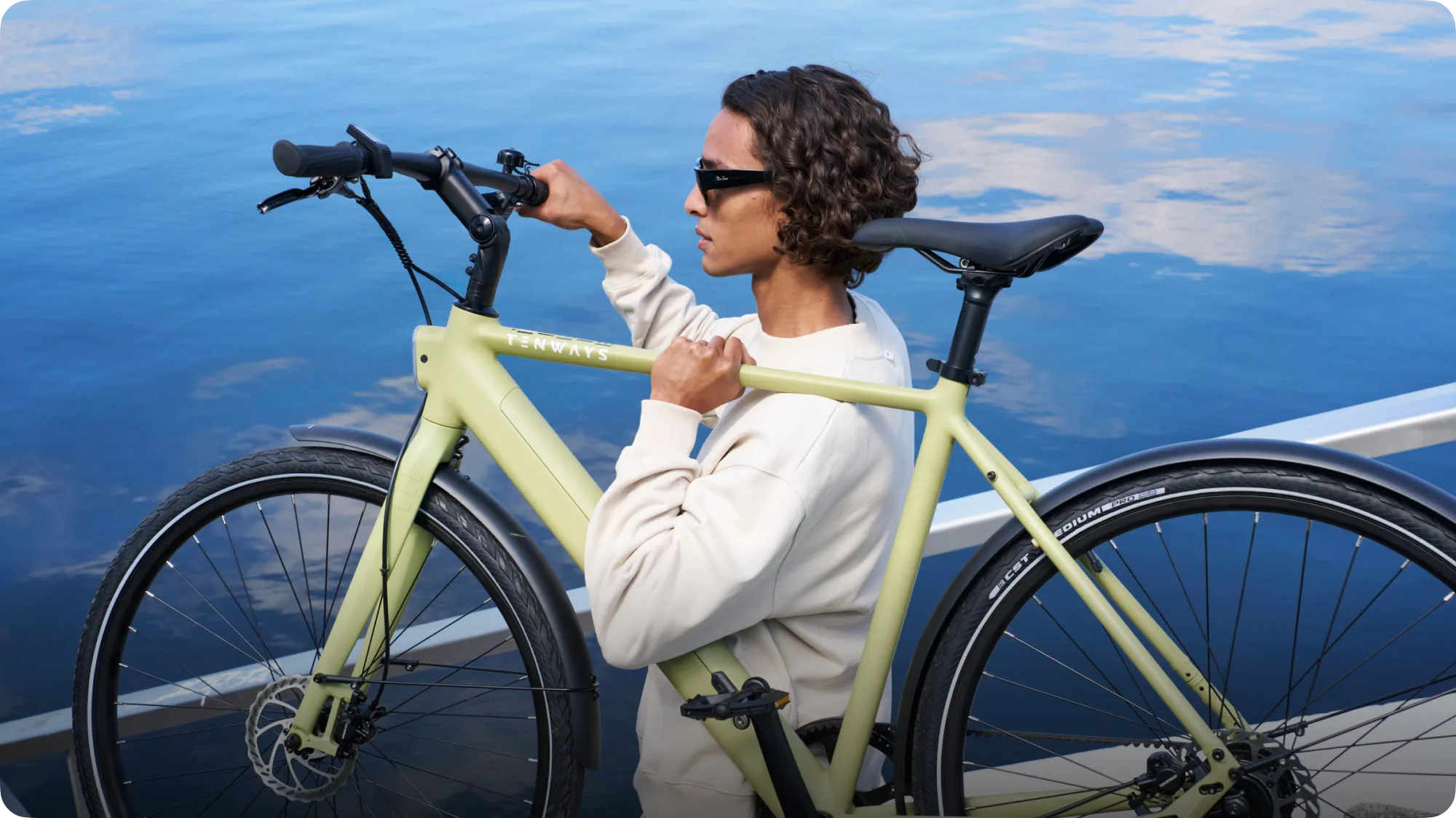 Ein Mann mit lockigen Haaren und Sonnenbrille trägt ein hellgrünes TENWAYS CGO600 Pro E-Bike in der Nähe von blauem Wasser.