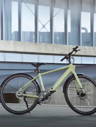 Eine Frau in grüner Kleidung steht neben einem hellgrünen TENWAYS CGO600 Pro E-Bike, das auf einer Betonfläche vor einem modernen Gebäude abgestellt ist.
