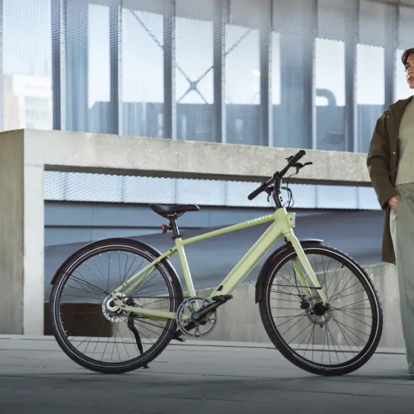 Eine Frau in grüner Kleidung steht neben einem hellgrünen TENWAYS CGO600 Pro E-Bike, das auf einer Betonfläche vor einem modernen Gebäude abgestellt ist.