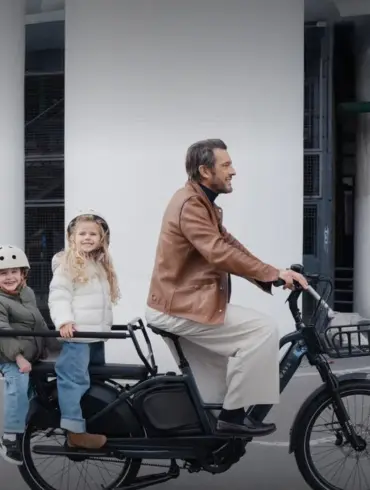 Ein Mann fährt ein schwarzes TENWAYS LONGTAIL DUO Lastenrad mit zwei Kindern auf der hinteren Sitzbank. Die Kinder tragen Helme und warme Jacken.