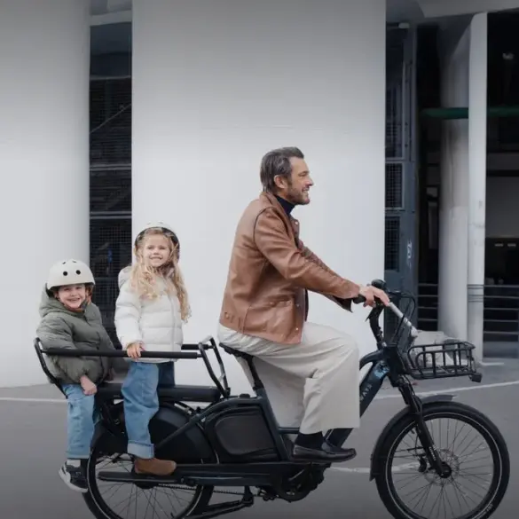 Ein Mann fährt ein schwarzes TENWAYS LONGTAIL DUO Lastenrad mit zwei Kindern auf der hinteren Sitzbank. Die Kinder tragen Helme und warme Jacken.