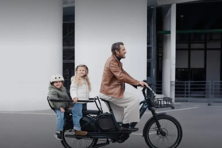 Ein Mann fährt ein schwarzes TENWAYS LONGTAIL DUO Lastenrad mit zwei Kindern auf der hinteren Sitzbank. Die Kinder tragen Helme und warme Jacken.