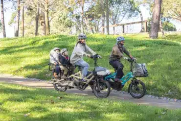 Familie, bestehend aus zwei Erwachsenen und zwei Kindern, fährt auf zwei kompakten Lasten-E-Bikes im Park.