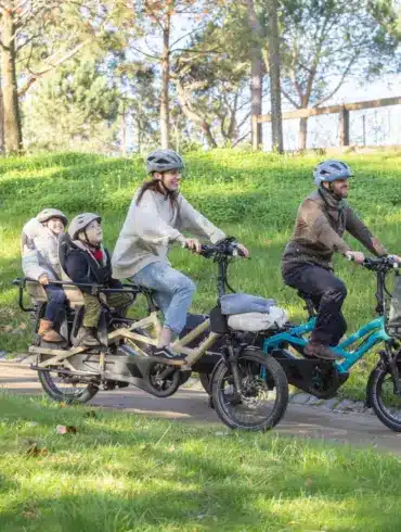 Familie, bestehend aus zwei Erwachsenen und zwei Kindern, fährt auf zwei kompakten Lasten-E-Bikes im Park.