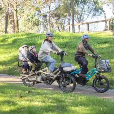 Familie, bestehend aus zwei Erwachsenen und zwei Kindern, fährt auf zwei kompakten Lasten-E-Bikes im Park.