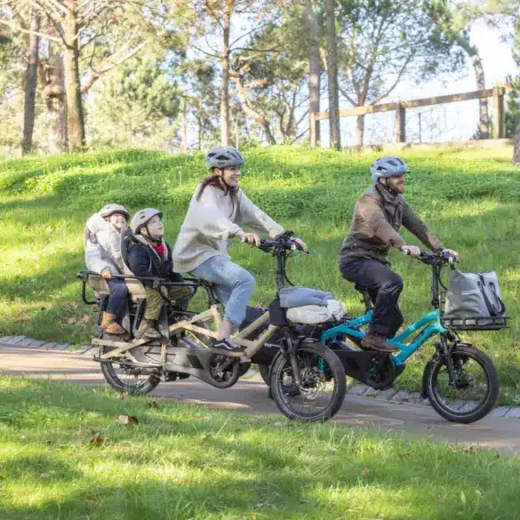 Familie, bestehend aus zwei Erwachsenen und zwei Kindern, fährt auf zwei kompakten Lasten-E-Bikes im Park.