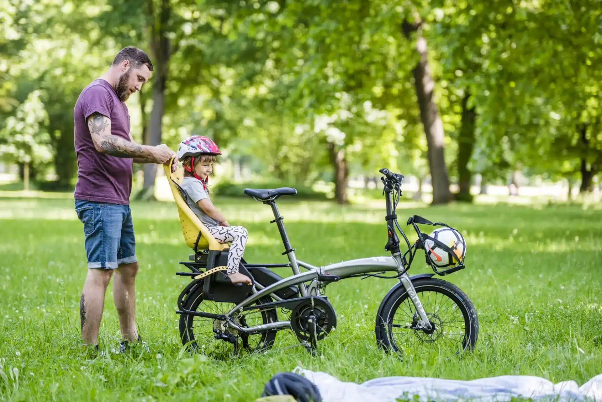 Vater stellt Kindersitz auf einem silbernen E-Faltrad in einer grünen Parklandschaft ein, während ein Kind darin sitzt.