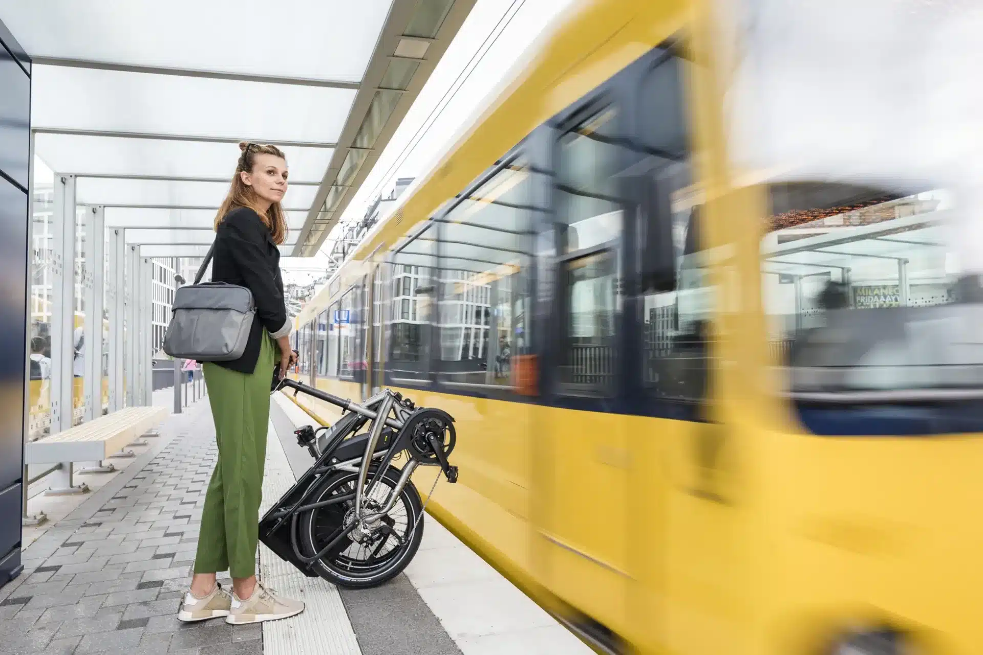 Frau mit schwarzem Blazer und grüner Hose steht mit einem zusammengeklappten silbernen Vektron S10 E-Bike auf einem Bahnsteig neben einer vorbeifahrenden gelben Straßenbahn.