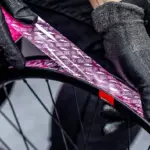Detailaufnahme von Händen mit schwarzen Muc-Off Handschuhen, die ein pinkes Muc-Off Tubeless Felgenband auf einer schwarzen Fahrradfelge anbringen.