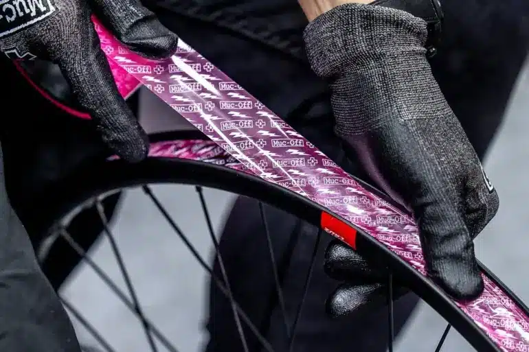 Detailaufnahme von Händen mit schwarzen Muc-Off Handschuhen, die ein pinkes Muc-Off Tubeless Felgenband auf einer schwarzen Fahrradfelge anbringen.