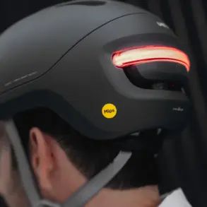 Ein schwarzer UNIT 1 NEON Urban Smart Helm wird von hinten auf dem Kopf einer Person getragen. Das markante rote LED-Rücklicht am Helm ist eingeschaltet und leuchtet hell. Ein gelber Mips-Aufkleber befindet sich seitlich am Helm. Der Helm hat ein modernes und zurückhaltendes Design.