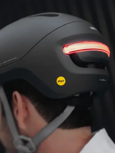Ein schwarzer UNIT 1 NEON Urban Smart Helm wird von hinten auf dem Kopf einer Person getragen. Das markante rote LED-Rücklicht am Helm ist eingeschaltet und leuchtet hell. Ein gelber Mips-Aufkleber befindet sich seitlich am Helm. Der Helm hat ein modernes und zurückhaltendes Design.