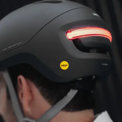 Ein schwarzer UNIT 1 NEON Urban Smart Helm wird von hinten auf dem Kopf einer Person getragen. Das markante rote LED-Rücklicht am Helm ist eingeschaltet und leuchtet hell. Ein gelber Mips-Aufkleber befindet sich seitlich am Helm. Der Helm hat ein modernes und zurückhaltendes Design.