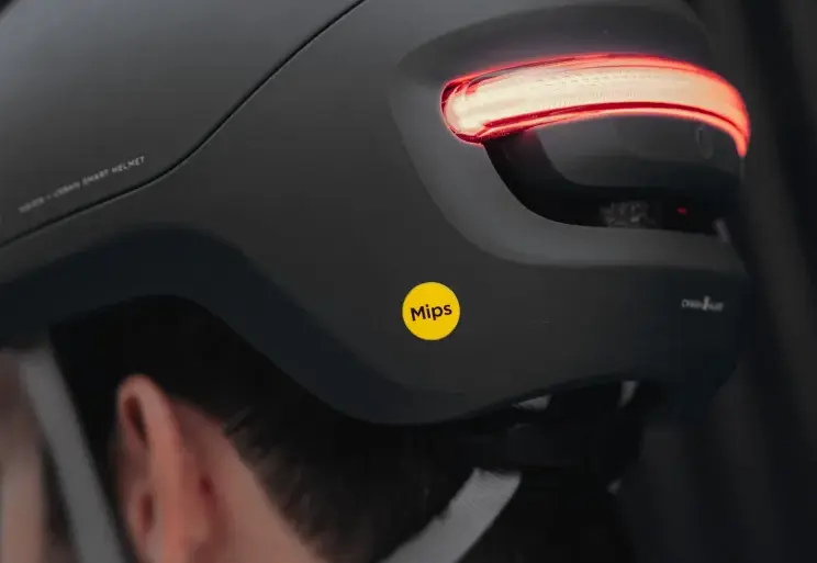 Ein schwarzer UNIT 1 NEON Urban Smart Helm wird von hinten auf dem Kopf einer Person getragen. Das markante rote LED-Rücklicht am Helm ist eingeschaltet und leuchtet hell. Ein gelber Mips-Aufkleber befindet sich seitlich am Helm. Der Helm hat ein modernes und zurückhaltendes Design.