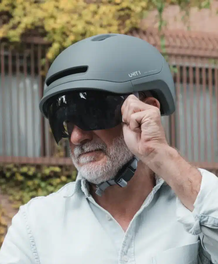Porträt eines älteren Mannes mit grauem Bart, der einen modernen grauen Fahrradhelm der Marke UNIT 1 auf dem Kopf trägt. Der Helm ist mit einem dunklen Visier ausgestattet, das er gerade mit seiner rechten Hand anpasst. Im Hintergrund ist eine grüne Hecke und ein Zaun zu sehen.