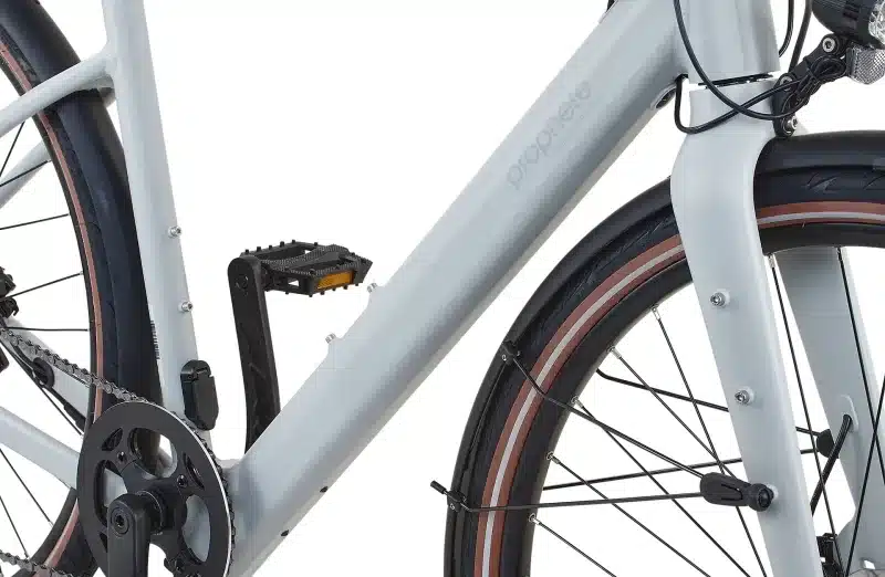 Detailaufnahme eines hellgrauen Prophete Urbanicer 2.9 E-Bikes mit Fokus auf den Rahmen, das Pedal und das Vorderrad mit Schutzblech.