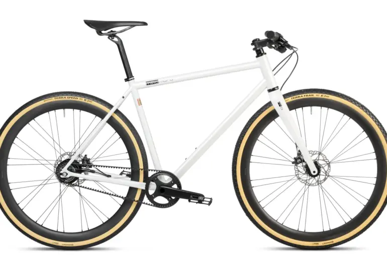 Ein weißes Gravel Bike mit Riemenantrieb und beigen Reifen, stehend vor weißem Hintergrund.