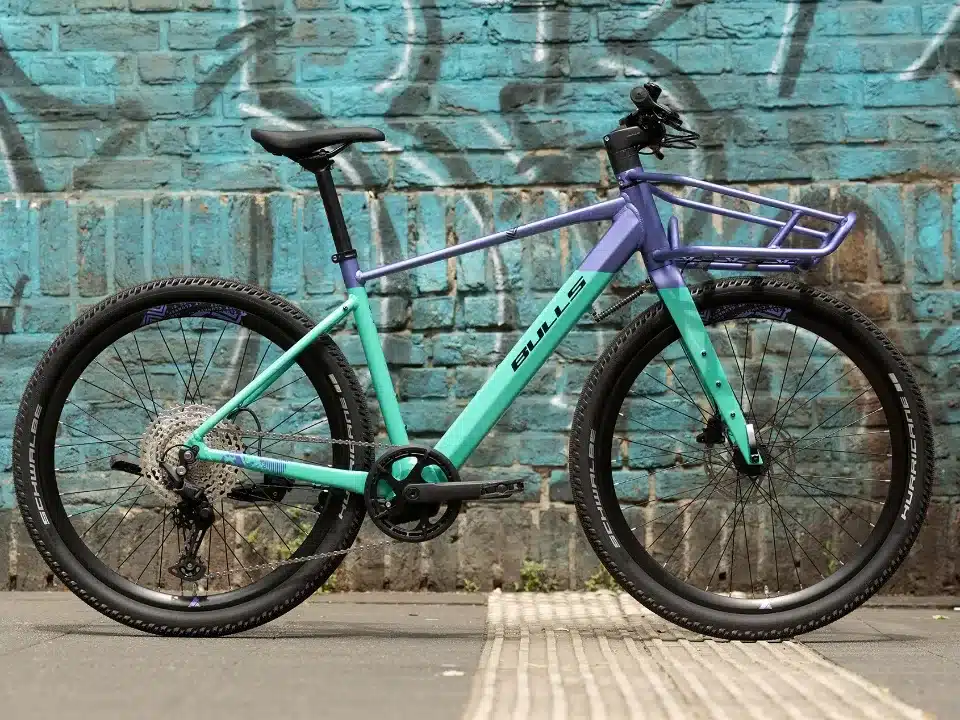 Bulls Clunker Evo E-Bike in turquoise und lila mit integriertem Frontgepäckträger, abgestellt vor einer bunten Ziegelmauer.