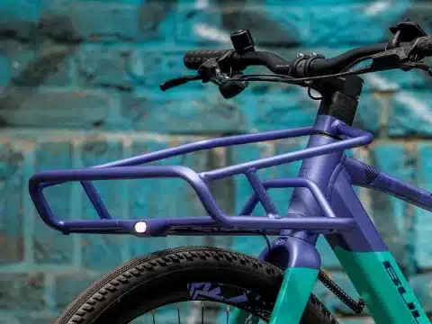 Vorderansicht des Bulls Clunker Evo E-Bikes mit integriertem, violettem Frontgepäckträger und Fahrradlicht, vor einer blauen Ziegelwand.